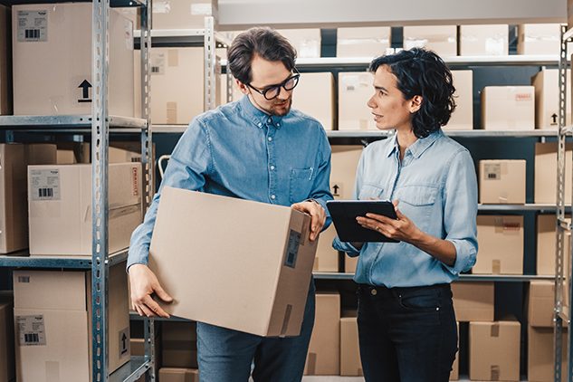 man and woman in warehouse with boxes look at tablet