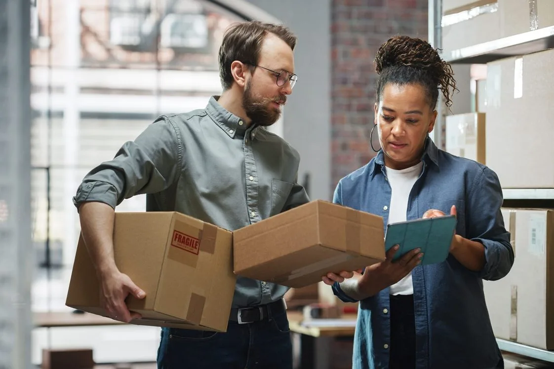 Inventory Manager Using Tablet Computer, Talking to a Worker Holding Two Cardboard Packages. They Discuss Customer Orders. Stock of Parcels with Products Ready for Shipment in the Background.