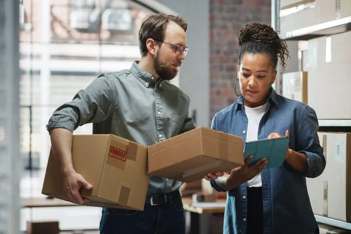 Inventory Manager Using Tablet Computer, Talking to a Worker Holding Two Cardboard Packages. They Discuss Customer Orders. Stock of Parcels with Products Ready for Shipment in the Background.