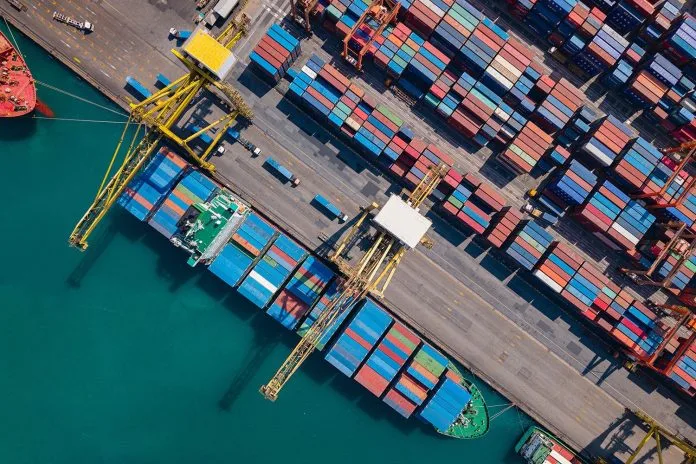 Drone shot of a bustling shipping port with colorful containers and giant cargo cranes beside bright turquoise ocean waters.